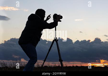 Ein Mädchen fotografiert den Sonnenuntergang von einem Stativ aus, das auf dem Dach eines Autos auf dem Feld steht und den Raum kopiert. Stockfoto