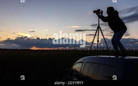 Ein Mädchen fotografiert den Sonnenuntergang von einem Stativ aus, das auf dem Dach eines Autos auf dem Feld steht und den Raum kopiert. Stockfoto