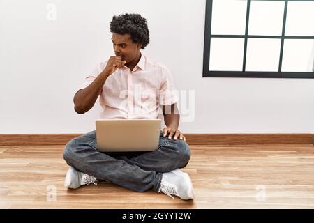 Afrikanischer Mann mit lockigen Haaren mit Laptop sitzt auf dem Boden Gefühl, sich unwohl und Husten als Symptom für Kälte oder Bronchitis. Gesundheitskonzept. Stockfoto