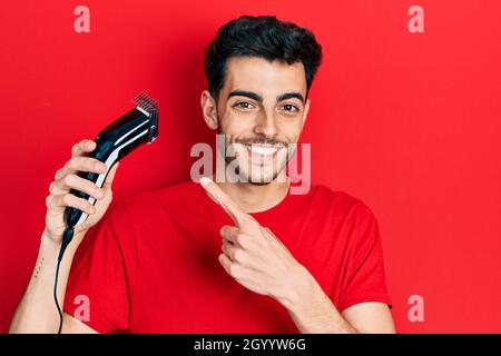 Junger hispanischer Mann lädt elektrische Rasiermaschine lächelnd glücklich zeigt mit Hand und Finger Stockfoto