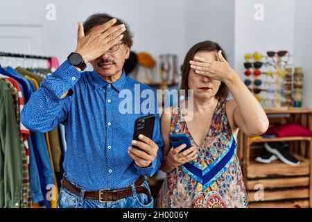 Interrassisches Paar im mittleren Alter im Einzelhandel mit Smartphone, das die Augen mit der Hand bedeckt, sieht ernst und traurig aus Stockfoto