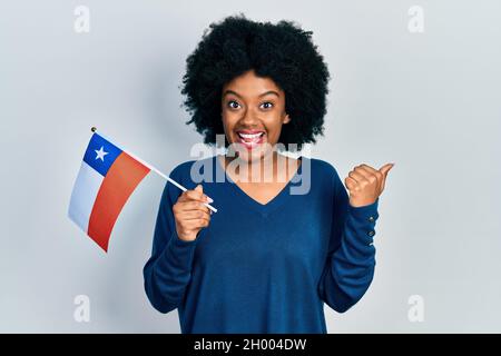 Junge afroamerikanische Frau, die die chilenische Flagge hält und den Daumen nach oben zur Seite zeigt, lächelt glücklich mit offenem Mund Stockfoto