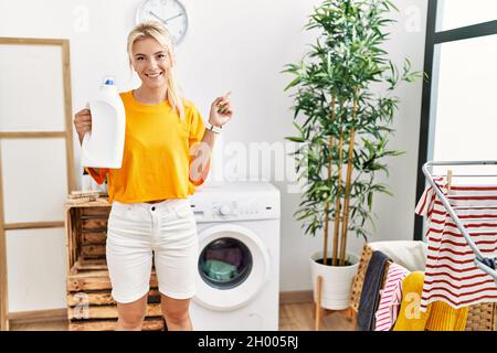 Junge kaukasische Frau, die ihre Wäsche waschen und die Waschmittelflasche lächelnd hält, zeigt glücklich mit Hand und Finger zur Seite Stockfoto