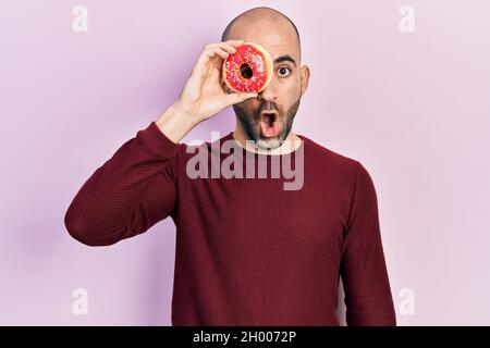 Junger kahler Mann, der einen leckeren bunten Donut auf dem Auge hält, erschreckt und überrascht mit offenem Mund für ein überraschendes, ungläubig gesichtes Gesicht Stockfoto