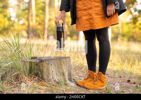 Weibliche Touristin in orangefarbenen Kleidern, die eine Thermoskanne auf einen Baumstumpf im Wald legt, um eine Kaffeepause zu machen und sich aufzuwärmen. Waldwandern in au Stockfoto