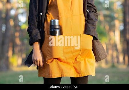 Weibliche Wanderung in einem orangefarbenen Kleid und mit einer Thermoskanne in der Tasche, die vor einem verschwommenen Waldhintergrund steht. Stockfoto
