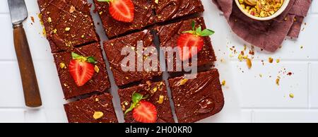 Schokoladenquadrate mit Pistazien und Erdbeeren auf weißem Papier auf hellem Hintergrund, Draufsicht, horizontale Komposition. Flache Lebensmittel. Stockfoto