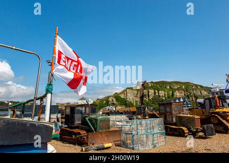 Die Flagge von St. George wird von Fischerbooten an der Küste von Hastings in East Sussex, England, mit der Standseilbahn in der Ferne überflogen Stockfoto