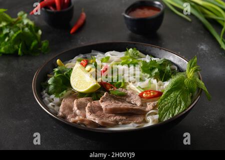 Pho Bo traditionelle Suppe mit Rindfleisch, Reisnudeln, Ingwer, Limette, Chilischote in einer Schüssel auf schwarzem Hintergrund. Nahaufnahme. Vietnamesische und asiatische Küche. Stockfoto