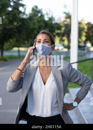 Hübsche junge Geschäftsfrau, die eine schützende Gesichtsmaske trägt und vor dem Büro mit dem Mobiltelefon spricht Stockfoto