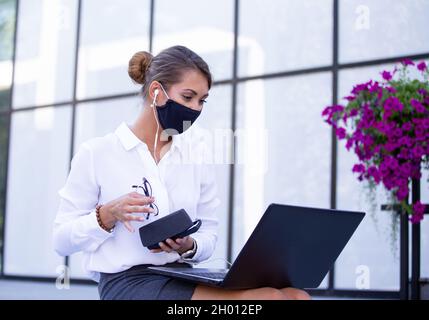 Eine hübsche junge Geschäftsfrau, die eine Schutzmaske trägt und vor dem Bürogebäude an einem Laptop arbeitet Stockfoto