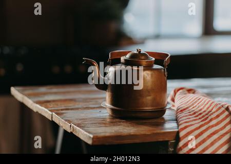 Horizontale Aufnahme der Aluminium-Teekane auf Holztisch vor verschwommenem Hintergrund gestreifte Serviette in der Nähe. Alter Kessel aus Bronze Stockfoto