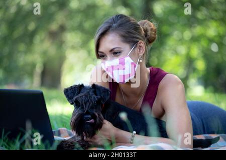 Geschäftsfrau mit Schutzmaske, die auf einem Laptop im Park mit Hund neben ihr arbeitet Stockfoto