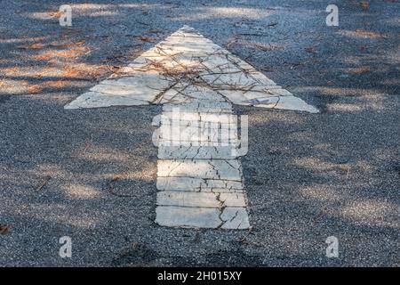 Großer weißer nach vorne gerichteter Pfeil auf dem nassen Asphalt im Schatten, der durch den Verkehr auf der Straße im Freien mit Sonnenlicht zerrissen und abgenutzt ist Stockfoto