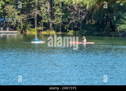 Zwei Männer fischen in Kajaks zusammen auf dem See in der Bucht entlang der Küste mit den Wäldern und dem schwimmenden Dock im Hintergrund auf einem sonnigen Stockfoto