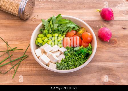 Vegane Poke-Schüssel mit Tofu, Grünkohl, gehacktem Schnittlauch, Edamame-Bohnen, gehackten Kirschtomaten, Rucola-Blätter und weißer Reis auf dem Takeaway-Behälter Stockfoto