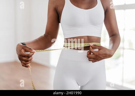 Gewichtsverlust Konzept. Nahaufnahme beschnittene Ansicht einer nicht recognozable fit afroamerikanische Dame, die ihre Taille mit gelbem Klebeband misst, verschwommener Hintergrund. Schlank Stockfoto