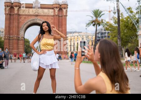Junge asiatische Reisende in trendigem Sommeroutfit posieren vor dem Triumphbogen, während die Freundin während der Sightseeing-Tour in Barcelona Fotos auf dem Smartphone fotografiert Stockfoto