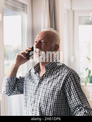 Aufgeregter alter Mann im Hemd mit weißen Haaren und Bart, der zu Hause neben dem Fenster telefoniert Stockfoto