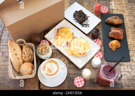 Brunch zum Mitnehmen mit Eiern benedict, verschiedenen Brötchen, Joghurt mit Nüssen, Marmelade und Schokoladencreme, Butter und verschiedenen Gebäckstücken mit Erdbeer-Smoothie Stockfoto