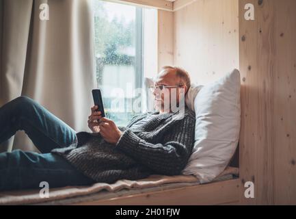 Lächelnder Mann mittleren Alters gekleidet warme Strickjacke, Jeans liegen auf einem gemütlichen Bett neben dem Fenster und mit einem modernen Smartphone. Lifestyle-Foto für den Alltag wi Stockfoto
