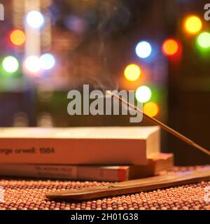 Entspannender Räucherstäbchen, der in einem Räuchergefäß mit Bokeh von Büchern und Hintergrundlichtern brennt. Stockfoto