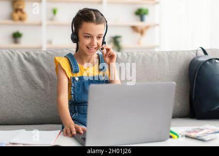 Kinder- Und Technologiekonzept. Porträt eines lächelnden Mädchens, das auf der Couch am Tisch sitzt und einen Laptop benutzt, das Headset trägt und berührt und im Mikrofon spricht Stockfoto