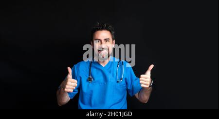 Ein Arzt in blauer Krankenhausuniform, dunkel und bärtig, mit einem Stethoskop um seinen Hals, lächelnd und mit einem Daumen nach oben, was auf Th hinweist Stockfoto
