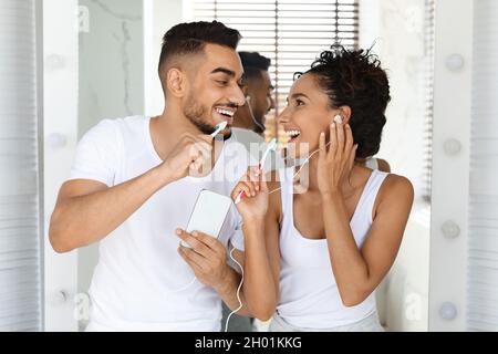 Glückliches Arabisches Paar Musik Auf Dem Smartphone Hören Und Singen, Während Man Im Badezimmer Zähne Putzt, Fröhliche Junge Ehepartner, Die Sich Die Kopfhörer Teilen Und Spaß Haben Stockfoto