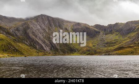 Sonnenlicht durchdringt die dunklen Wolken über dem Lake Idwal im Snowdonia National Park und beleuchtet die Bergseite und die Regenwasserstraßen, die herunterfließen. Stockfoto