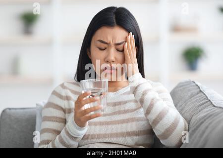 Kopfschmerz-Konzept. Kranke Junge Asiatische Frau, Die Zu Hause An Migräne Leidet, Kranke Koreanische Frau, Die Ein Glas Wasser Hält, Den Tempel Berührt Und Stirnrunzeln, Fühlt Stockfoto