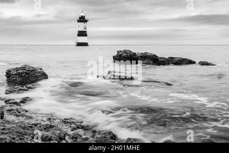 Schwarz-weißes Bild von Wellen, die um den Penmon Lighthouse und die zerklüfteten Felsen im Vordergrund herumschlagen. Auf der Anglesey Küste in Nort Stockfoto