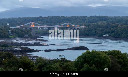 Die Menai Suspension Bridge wurde an einem Abend im Oktober 2021 über die Menai Strait an der Küste von Nordwales hinweg gesehen, als das Licht verblasst. Stockfoto