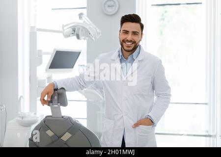Hübscher kaukasischer junger Arzt posiert in der neuesten Zahnklinik, Kopierraum. Glücklicher bärtiger männlicher Zahnarzt begrüßt Patienten oder lädt zum Besuch von Kleie ein Stockfoto