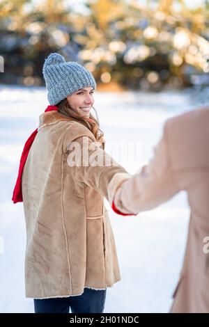 Lächelnde junge hübsche Dame, die die männliche Hand zieht, Porträt einer fröhlichen Frau, die mit ihrem Freund gemeinsam den ersten Winterschnee im Wald genießt und dabei die Kleinen verbringt Stockfoto