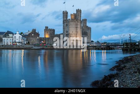 Caernarfon Castle dominiert bei Dämmerung im Oktober 2021 die Skyline und das Wasser an der Küste von Nordwales. Stockfoto