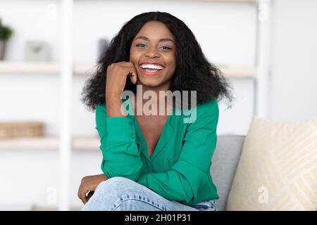 Fröhliche attraktive junge schwarze Dame posiert allein zu Hause, trägt stilvolles Casual-Outfit, sitzt auf der Couch und lächelt an der Kamera, gemütliches Wohnzimmer int Stockfoto