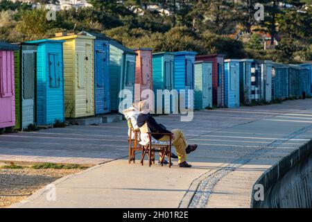 Ein älteres Paar mittleren Alters sitzt auf Stühlen am Meer neben einer Reihe von bunten Strandhütten in bembridge oder St. helens auf der Insel wight uk. Stockfoto