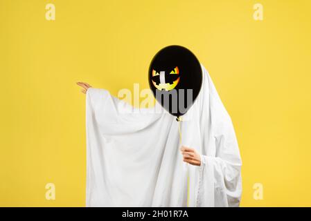 Halloween-Konzept. Mädchen in einem lustigen Geisterkostüm mit einem schwarzen Ballon mit Jack Laterne oder gruseligem Kürbisgesicht Stockfoto