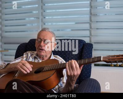 Glücklicher alter Mann, der zu Hause Akustikgitarre spielt und in seinem Sessel singt Stockfoto