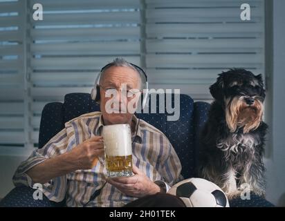 Älterer Mann mit Hund, der zu Hause im Sessel sitzt, Fußball und Bier hält und fernsieht Stockfoto