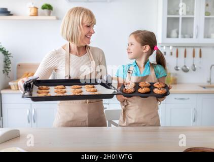 Kochen Lernen. Porträt einer lächelnden, reifen Großmutter und Enkelin, die leckere frisch gebackene Plätzchen und Muffins auf dem Tablett zeigen, während sie darauf stehen Stockfoto