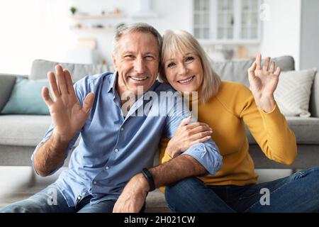 Hallo. Fröhliches Älteres Paar Winkt Die Hände Lächelnd Zur Kamera, Posiert Auf Dem Sofa Und Umarmt Sich Zu Hause. Glückliche Ehe Und Beziehung Im Hohen Alter. Stockfoto