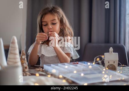 Nettes kleines Mädchen mit lockigen Haaren basteln und dekorieren Weihnachtsbaum Kegel mit Knöpfen. Stockfoto