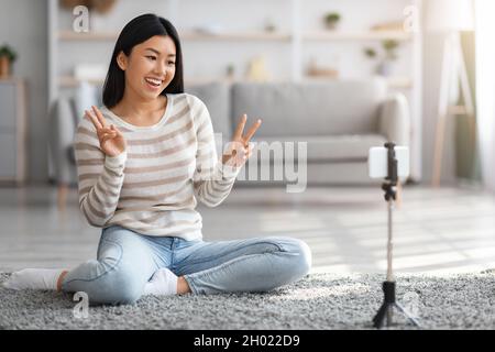 Glückliche junge asiatische Frau, die zu Hause Selfie macht oder Video-Blog einfängt, fröhliche Millennial Koreanische Frau, die Smartphone auf dem Stativ benutzt, gesturing Peace A Stockfoto