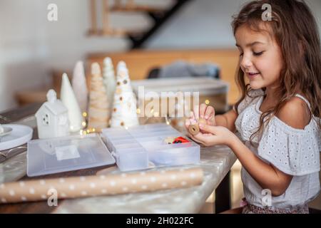 Nettes kleines Mädchen mit lockigen Haaren basteln und dekorieren Weihnachtsbaum Kegel mit Knöpfen. Stockfoto