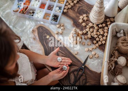 Nettes kleines Mädchen mit lockigen Haaren basteln und dekorieren Weihnachtsbaum Kegel mit Knöpfen. Stockfoto