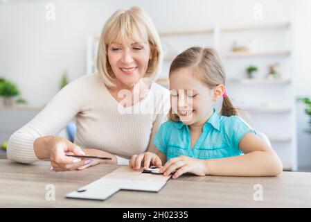 Liebevolle lächelnde reife Frau sitzt am Tisch mit kleinen Mädchen in Spiel verlobt Puzzle zusammen, glückliche Großmutter spielen Spaß mit Präs Stockfoto