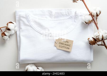 T-Shirt aus Bio-Baumwolle mit Baumwollblumen auf weißem Hintergrund. Flach liegend, Draufsicht. Öko-Bekleidung, nachhaltiger Lebensstil, Modekonzept. Stockfoto