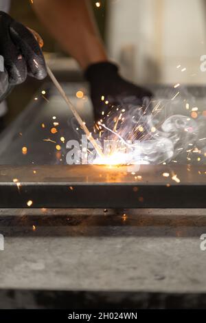Nahaufnahme der Hände einer Person, die schwarze Schutzhandschuhe trägt, während sie ein Metallschweißgerät verwendet, Funken von der Maschine, manuelle Maschinenarbeiten Stockfoto
